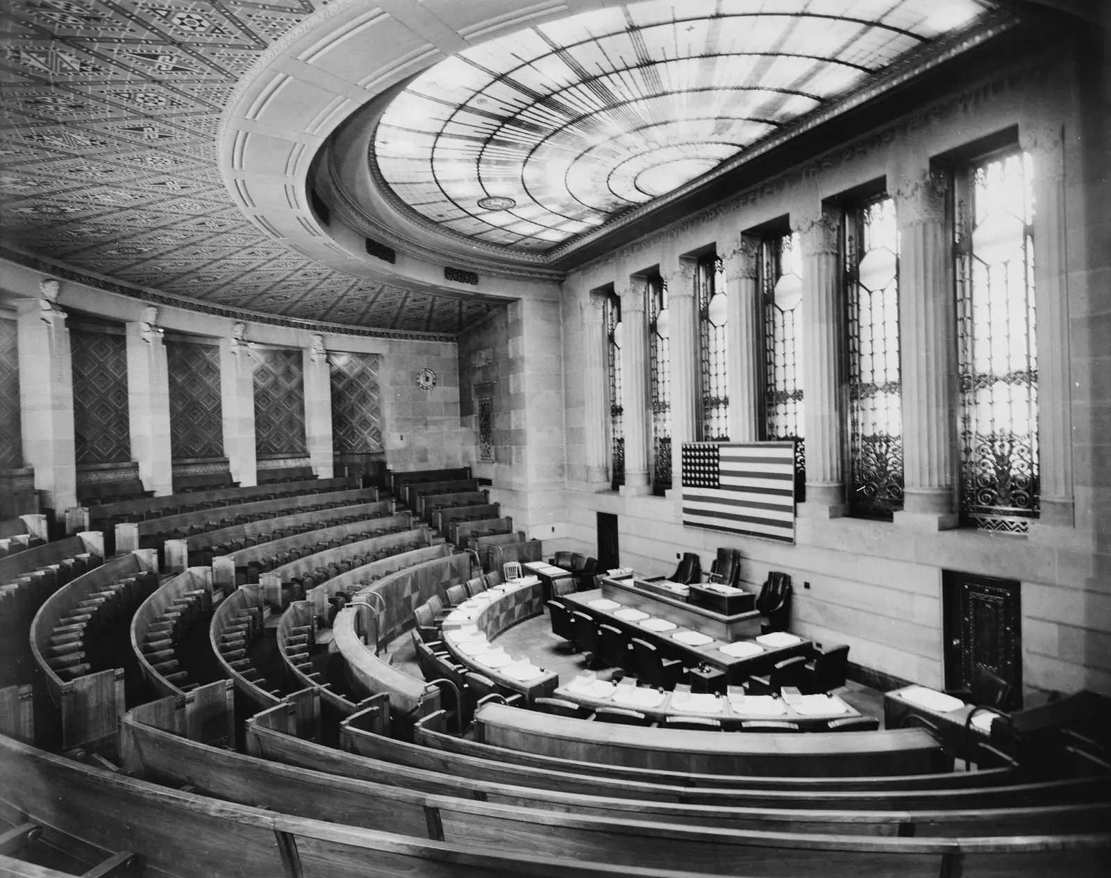 An empty city council chamber before a meeting: curved rows of seats, council desks with papers set out, the flag on the wall, and a glass skylight overhead.